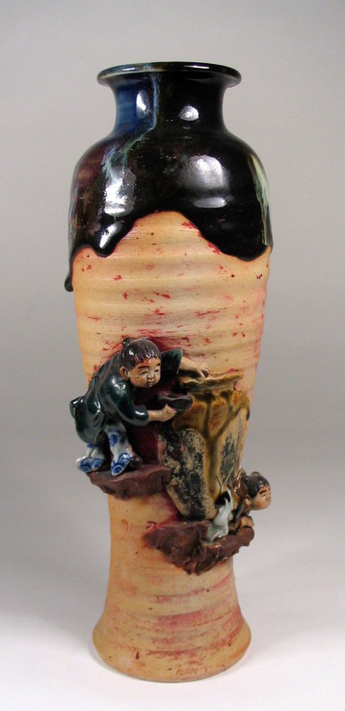 DESCRIPTION:  A tall Japanese Sumida Gawa (Sumida River) vase featuring two hand molded children in high relief. The rim and neck of the vase are covered in a rich, dark flambé glaze that runs down the shoulders onto the ribbed body.  Each of the children are perched on a ledge above and below a large, flat vase. The child at the top puts something in, while at the bottom of their vase we see a crack where water pours out.  Signed "Ryosai" on small blue & white cartouche; excellent condition with no chips or repairs.  DIMENSIONS:  12" high (30.5 cm) x 4 1/4" diameter (10.8 cm).  

<P>ABOUT SUMIDA EARTHENWARE POTTERY: This charming and highly collectable studio pottery originates from the Sumida River area in Japan (near Tokyo).  Produced primarily for export from the late 19th Century through the early 20th Century, this form of pottery is valued for the uniqueness of its handmade relief figures of people and animals engaged in various activities, often humorous or depicting scenes from folklore.  The tops are usually covered with a thick glaze in earth tones while the bodies of the pieces have an applied “cold paint” (usually red).  
<div id='rater_target1254611'></div>
