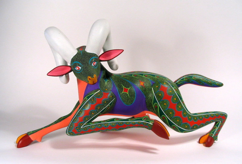 DESCRIPTION:  This dynamic and colorful Alebrije goat (or ram) is carved as if in a full run, its long front leg folded under its body in mid-stride.  Two huge white horns curl from the top of its head to its ears.  His green speckled body is accented with vibrant red "stripes" running down his back, tail and legs, while a purple design covers his belly.  This large, delightful animal is signed on the underside, "Narcisco Gonzalez Ramirez, Arrazola, Oaxaca Oax., Mexico."  Excellent condition.  DIMENSIONS:  19" long x 9.75" high.   

<p>CULTURAL BACKGROUND:  Oaxacan woodcarvings, also known as Alebrijes, have been a tradition for generations in several small villages outside the capital of Oaxaca, Mexico. It has only been in the past 40 years that these beautifully carved and painted figures have become popular, and are now collected worldwide. Using rudimentary tools, these Mexican artists create fascinating figures out of fresh-cut copal wood. After drying and sanding, the carvings are meticulously painted with intricate patterns and vibrant colors, limited only by each artist’s imagination.  Collectors highly value the signed pieces; however, the Alebrije may have had many hands contribute to its making.  Often the person who signs the alebrije is the person who is the most well known in the family or the workshop. A son may carve an alebrije in his father’s workshop, a grandson may sand it and a daughter may paint it. But if the father is the most well known carver, it is signed with his name. 
<div id='rater_target1309026'></div>
