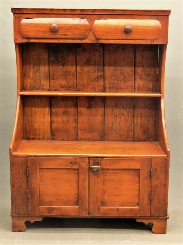 DESCRIPTION:  This early to mid 19th c. high-back pine bucket-bench, or cupboard, is a graceful and highly unusual piece of American country & primitive furniture. With shaker-like sensibility, this utilitarian piece would have been at home in the foyer where you could sit while taking off snowy boots, or in the kitchen used as storage for crocks of grain and other staples, or on the covered porch by the entry where you could place groceries or packages while opening the front door. 
<p>This bucket bench was created with graceful, curving sides enclosing two drawers at the top, a mid-height narrow shelf, and a bench with a two-door storage compartment beneath, all supported on attractive, carved bracket feet.  CONDITION: Age appropriate wear, scratches and stains; both top drawers have “old damage” slivers of wood missing on the front top left corners (see photos). DIMENSIONS: 42" wide x 18" deep x 5' high.
<div id='rater_target1370789'></div>
