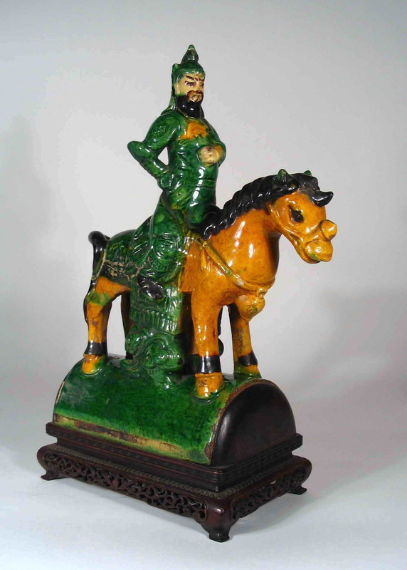 DESCRIPTION:  A beautifully mounted Chinese roof tile of a Ming warrior seated upon a horse.  The bearded soldier, his right hand on his hip, sits erect upon his amber horse which stands on the curved tile base. This earthenware tile has been decorated in a sancai glaze of predominately green and amber colors. A beautifully carved custom rosewood base has been made for this large tile, the base itself probably being over 100 years old.  CONDITION: Quite good condition with the usual wear one would expect from Ming Dynasty piece including small chips.  The stand has been drilled with a hole on one side, presumable for using as a lamp.  Ming Dynasty, 1368 – 1644.  DIMENSIONS:   16 ½” tall with stand (42 cm) x 11” wide (28 cm).  
<p>HISTORICAL BACKGROUND: During the Ming and Qing Dynasties, glazed sculptural tiles became a popular decorative devise used extensively on temples, altars, imperial palaces and gardens, and are considered one of the hallmarks of classical Chinese architecture. Eaves and entryways were decorated with vibrant sculptural tiles that served both decorative and religious purposes. On temples and palaces, representations of mounted warriors and snarling dragons were meant to ward off evildoers of both the physical and spiritual kind. 

<p>The term “Sancai” literally translates: “three colors”, and is usually applied to Tang dynasty earthenwares, which are coated with slip and then decorated with lead-fluxed glazes. The three most common colors, from which the name derives, are cream, amber and green. The sancai palette was revived in later periods, and was used on ceramics in the Ming and Qing dynasties. 
<div id='rater_target1273089'></div>
