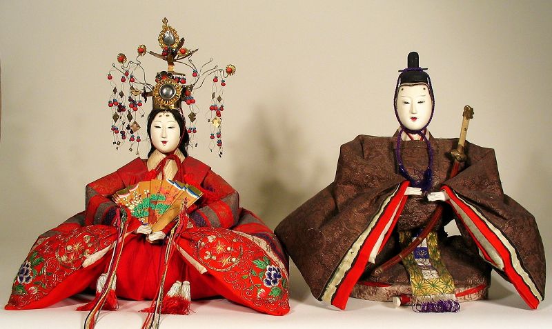 DESCRIPTION: A pair of large Japanese Emperor and Empress dolls, or kokin-bina, presented in formal court dress. The empress’ attire is quite elaborate, using fine brocade silks with hand embroidered flowers. In her hands she holds a painted fan and is crowned with an elaborate headdress representing a bird with long hanging beads attached. Both dolls have gofun faces and hands, which are in excellent condition. The accessories include the empress’ crown and tasseled fan, and the emperor’s long sword with shagreen tsuka (handle) and traditional black hat. Meiji period, c. 1868-1912. 
<p>CONDITION: Fabrics are bright and in generally fair condition for both dolls; there is some scattered fragility with some fabric including the cummerbund fabric on the emperor’s back. DIMENSIONS: With accessories, the emperor is 12” high (30.5.cm); empress is 13” high (33 cm).   
<p>HISTORY: Every year on March 3rd, Japan celebrates The Doll Festival (Hina Matsuri), and until recently, this date was also Girl’s Day. On this day, families set up a special step-altar on which to arrange their dolls. They would decorate this altar with boughs of peach blossoms and make offerings to the hina dolls of rice cakes (mocha), either flavored with a wild herb or colored and cut into festive diamond shapes. 

<div id='rater_target1369695'></div>
