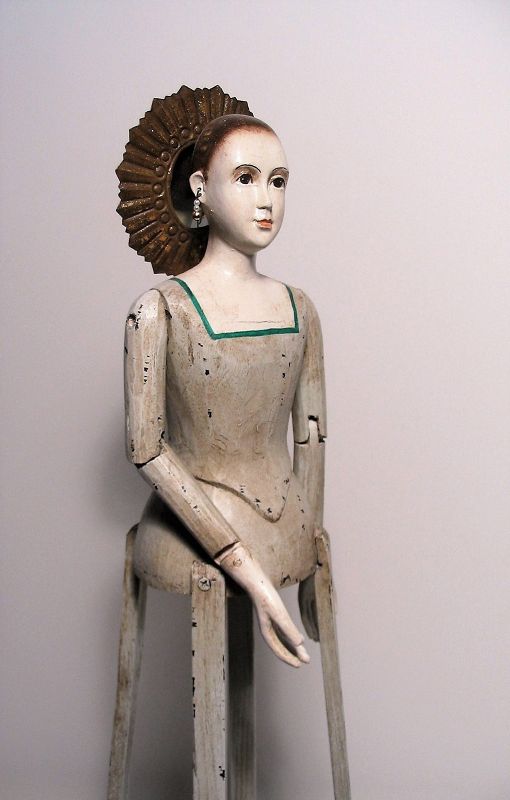 DESCRIPTION:  An elegant and stately santos cage doll (a Virgin Mary bastidor), with grey bodice and hand carved, delicate wooden face in sacred, peaceful expression, framed by a radiant gilt-tin halo. Her arms and hands are articulated from the shoulder; four wooden slats form the frame.  Good condition. DIMENSIONS: 24” high; 6 7/8” diameter base.
<div id='rater_target1387995'></div>

