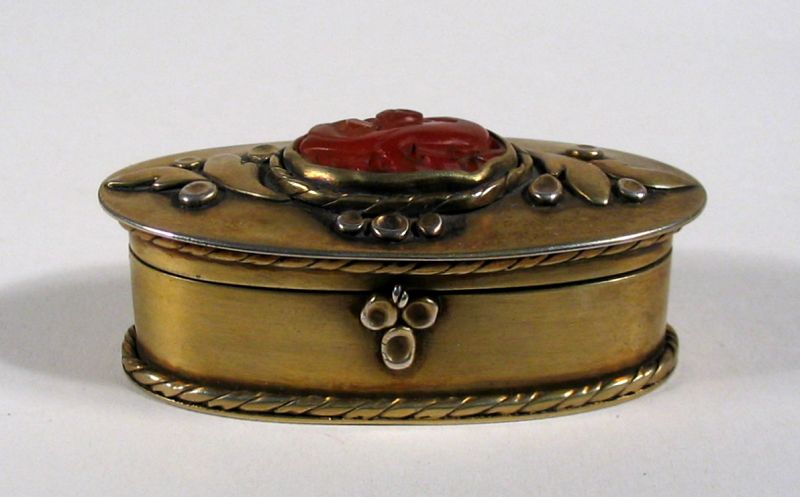 DESCRIPTION: An attractive oval brass box, the hinged lid decorated with a carved carnelian stone at the center with raised brass leaves on either side. Signed at the interior lid, such a box might have been used for snuff, jewelry, cosmetics or medicines. This attractive little box is in excellent condition and dates from the first quarter of the 20th C. DIMENSIONS: 2 ¼” long (5.7 cm) x 1 1/8” wide (2.9 cm) x 1” high (2.5 cm). 

<div id='rater_target1418151'></div>
